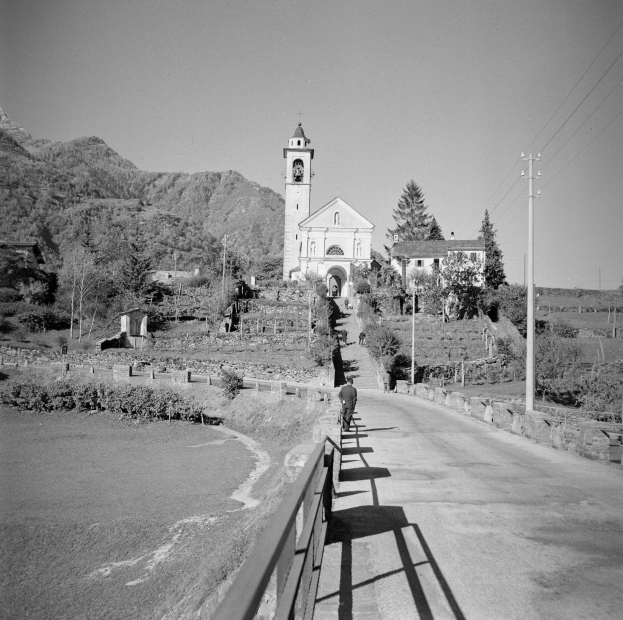 Schwarz-weißes Foto einer Person, die ein Fahrrad auf einer Straße mit Geländern fährt, umgeben von Bäumen, Gebäuden, Polen, Drähten, Hügeln und einem klaren Himmel.