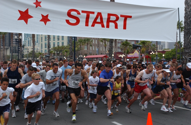 Gruppe von Läufern bei einem Marathon an einer Verkehrsinsel vorbei, mit einem Banner und Bäumen im Hintergrund unter einem klaren blauen Himmel.