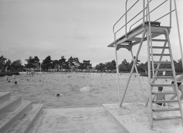 Schwarzes und weißes Foto von Menschen, die im Meer an einem Strand schwimmen, mit einem Lebensrettungsturm auf der rechten Seite mit einer Treppe, die hinaufführt, Bäumen und Pfählen im Hintergrund unter einem klaren Himmel.
