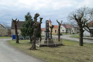 Ein kleines Dorf mit einem Friedhof in der Mitte, umgeben von Bäumen, Gras, einem Zaun, einer Straße, Fahrzeugen, Mülltonnen und Gebäuden mit Fenstern unter einem bewölkten Himmel, mit einem Gedenkstein mit eingravierter Inschrift auf dem Boden.