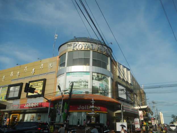 Eine belebte Stadtstraße mit fahrenden Fahrzeugen, Fußgängern auf dem Gehweg und einem großen Gebäude mit der Aufschrift "Königlicher Einkaufsbummel" im Hintergrund, umgeben von parkenden Autos, Bäumen, Strommasten und Drähten unter dem Himmel.