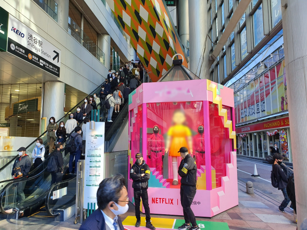 Eine Gruppe von Menschen steht vor einem Gebäude mit Glaswänden und Texttafeln, einige tragen Masken, während eine Rolltreppe mit Menschen daneben aufsteigt; ein pinker Kasten in der Mitte zeigt Schaufensterpuppen und trägt 'Netflix' neue Netflix-Bühne in Tokio'.