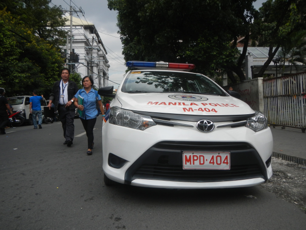 Polizeifahrzeug mit sichtbarem Nummernschild und Text am Straßenrand geparkt, einige Personen auf dem Gehweg, eine haltend ein Objekt, Geländer und Wand rechts, Bäume, Gebäude, Pfosten, Drähte und Himmel im Hintergrund.