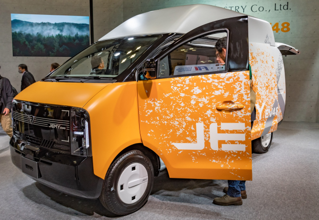 Orangefarbener und weißer Elektrovan auf einer Ausstellung, umgeben von Menschen in bunter Kleidung, mit einer Wandmontage und Text im Hintergrund.