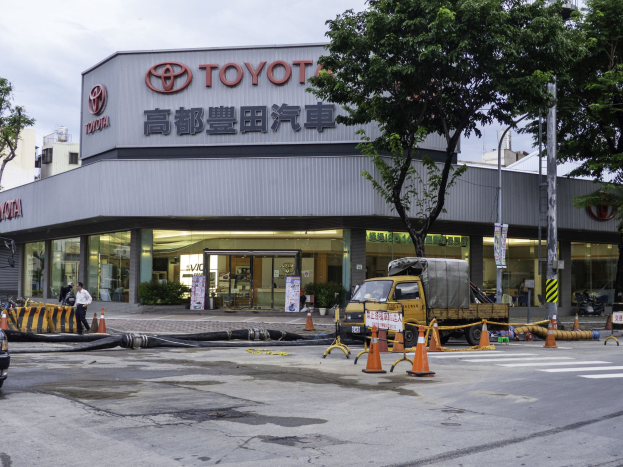 Außenansicht einer Toyota-Niederlassung in Tokio, Japan, mit Fahrzeugen, Verkehrskegeln, Rohren, Menschen, Bäumen, Polen, Plakaten, Gebäuden und einem bewölkten Himmel.