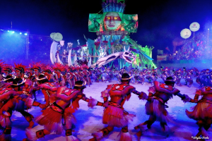 Eine Gruppe von Menschen in bunten Kostümen tanzt auf einer Bühne vor einem sitzenden Publikum, mit Lichtern, Polen und Ballons, und ein Bildschirm zeigt eine Person im Hintergrund, während des Karnevals in Rio de Janeiro 2014.