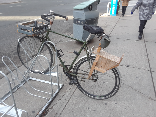 Ein grünes und braunes Fahrrad mit angebrachten Gegenständen steht an der Seite einer Straße neben einem Müllcontainer, mit Fahrzeugen auf der Straße und Menschen auf dem Gehweg im Hintergrund.