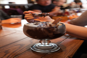 Eine Gruppe von Menschen sitzt an einem Tisch mit einem Glas Schokoladenpudding in der Mitte, eine Person im Vordergrund isst mit einem Löffel, Hintergrund leicht unscharf.