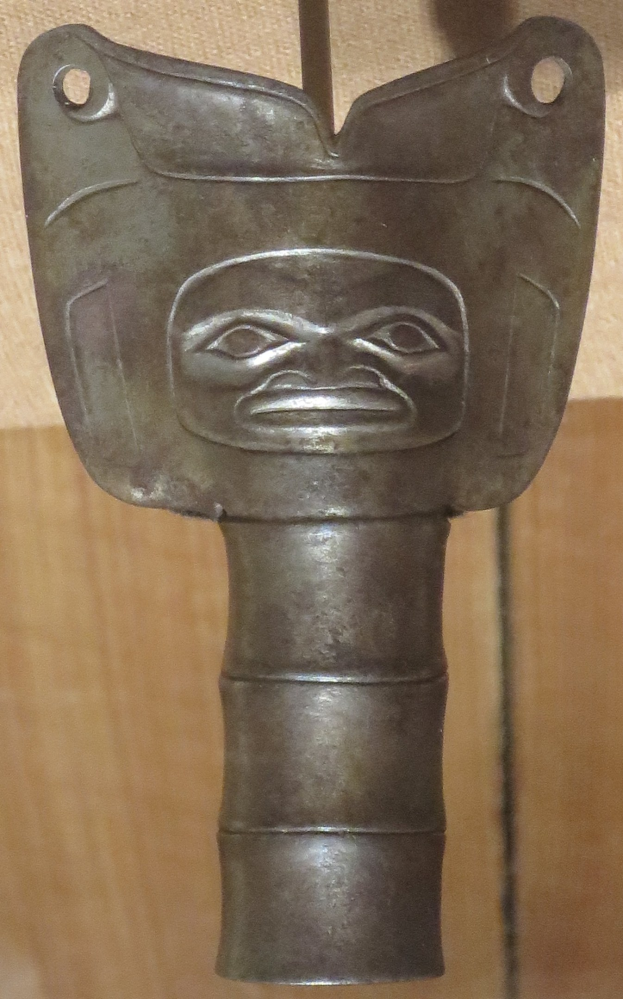 Nahaufnahme eines Metalldolches mit einer geschnitzten Gesichtsfläche darauf, das auf einem Holztisch gegen eine hölzerne Wand gestellt ist.