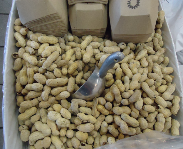 Eine Schachtel Erdnüsse auf einem Tisch mit einer Plastikabdeckung und einem Löffel daneben, umgeben von anderen verstreuten Erdnuss-Schachteln.