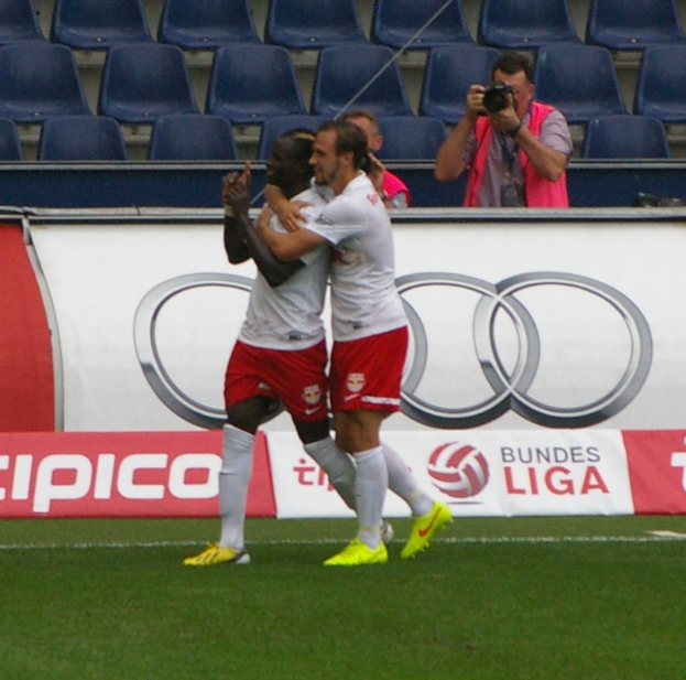 Zwei Fussballspieler feiern ein Tor auf einem Feld, umgeben von einer Gruppe von Menschen, mit einer Person, die eine Kamera hält, einem Banner, Stühlen und einer Wand im Hintergrund.
