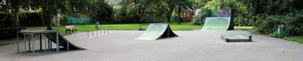 Ein Park mit einer zentralen Skaterrampe, umgeben von Bänken, Geländern, Gras, Pflanzen, Bäumen und Gebäuden im Hintergrund.