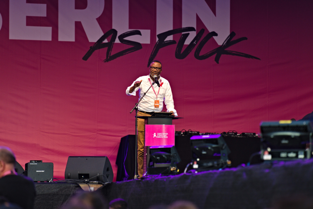 Mann in weißem Hemd und braunen Hosen, der in ein Mikrofon spricht, vor einer pinken 'Berlin as Fuck'-Flagge auf der Bühne.
