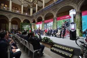 Eine Gruppe von Menschen sitzt in Stühlen und schaut auf einen Mann auf einer Bühne, der ein Mikrofon hält, mit Fahrrädern auf der rechten Seite und verschiedenen Elementen wie Plakaten, Pflanzen, Pfeilern, Geländern, Lichtern, Fenstern und einer Wand im Hintergrund.