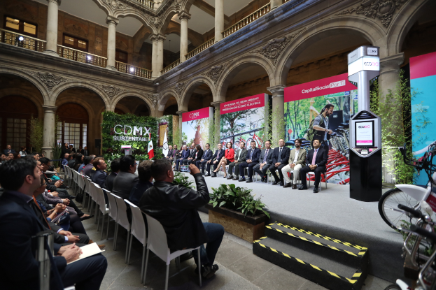 Eine Gruppe von Menschen sitzt in Stühlen und schaut auf einen Mann auf einer Bühne, der ein Mikrofon hält, mit Fahrrädern auf der rechten Seite und verschiedenen Elementen wie Plakaten, Pflanzen, Pfeilern, Geländern, Lichtern, Fenstern und einer Wand im Hintergrund.