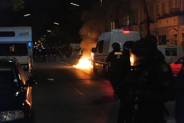 Polizisten stehen vor einem Feuer auf einer Straße bei Nacht mit Fahrzeugen und Gebäuden im Hintergrund.