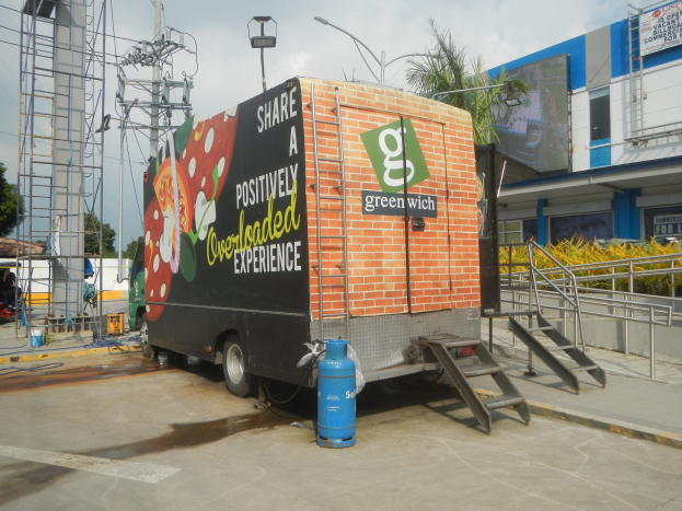 Ein großer Lkw vor einem Gebäude mit einer Tafel, auf der "Teilen Sie eine positiv überladene Erfahrung" steht, mit Stufen, einem Zylinder auf dem Boden, Geländern, Strommasten, Bäumen und einem bewölkten Himmel.
