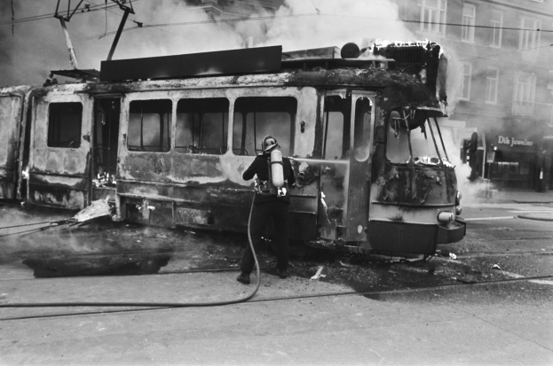 Schwarzes und wei├čes Bild eines Mannes in einem Helm, der eine Pfeife h├Ąlt und neben einer ausgebrannten Stra├čenbahn auf einer Stra├če steht, mit Rauch und einem Gebäude im Hintergrund.