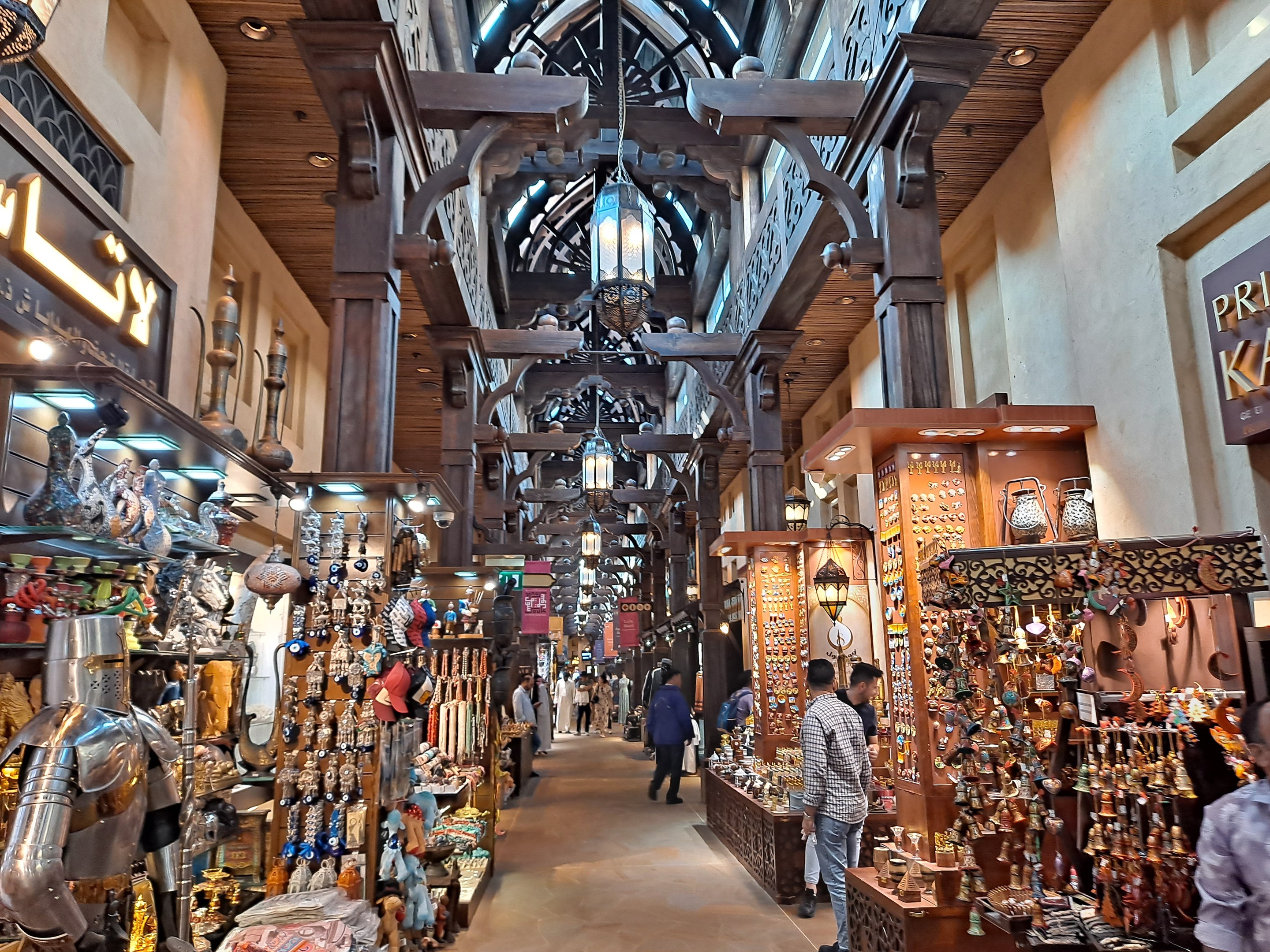 Ein geschäftiges Dubai-Markt mit Menschen, die zwischen Gestellen mit Waren hindurchgehen, Wänden mit Texttafeln und einer Decke mit warmem Licht.