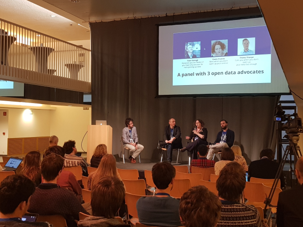 Eine Gruppe von Menschen sitzt auf Stühlen vor einem Projektionsschirm, mit einem Laptop auf einem Tisch, einer Kamera auf einem Stativ rechts daneben und Geländern, Lampen und einer Wand im Hintergrund während einer Podiumsdiskussion über Open-Data-Aktivisten.