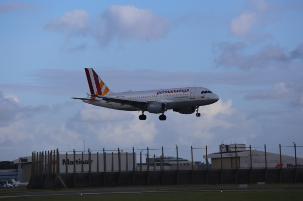 Germanwings Airbus A320-200 startet von Frankfurt Airport mit Gras darunter, einem Zaun im Vordergrund, Gebäuden und Bäumen im Hintergrund und einer bewölkten Himmel.