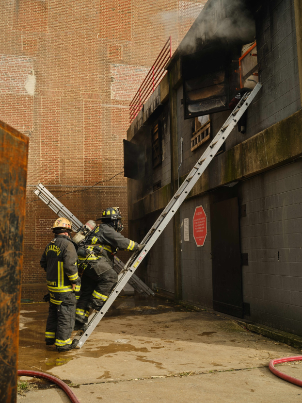 Feuerwehrleute mit Helmen klettern eine Leiter zu einem raucherfüllten Gebäude hoch, mit einem Metallobjekt links und Rohren auf dem Boden darunter.