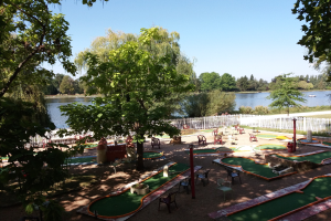 Minigolf-Platz in einem Park mit grünem Gras, Bäumen, einem Zaun, verstreuten Stühlen und Tischen, einem Gewässer mit einem Boot und sichtbarem Himmel im Hintergrund.