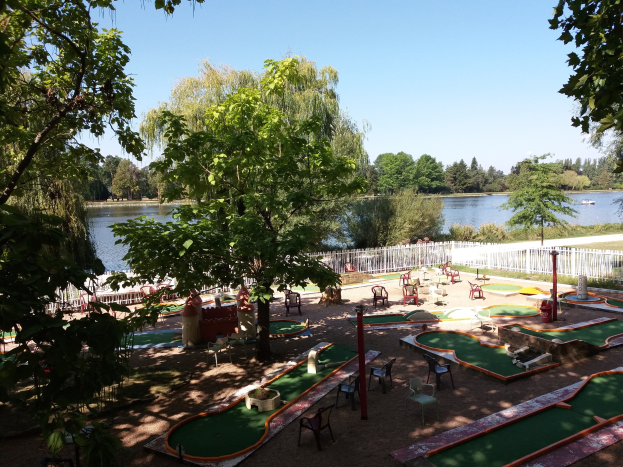 Minigolf-Platz in einem Park mit grünem Gras, Bäumen, einem Zaun, verstreuten Stühlen und Tischen, einem Gewässer mit einem Boot und sichtbarem Himmel im Hintergrund.