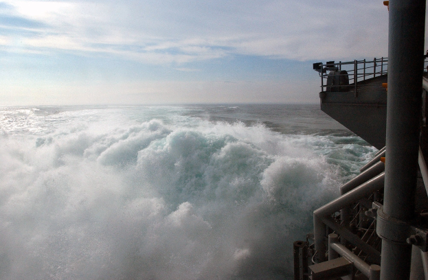 Große Welle, die über das Deck eines Schiffs mit Relingen und Pfählen auf der rechten Seite kracht, unter einem wolkenfüllten dramatischen Himmel.