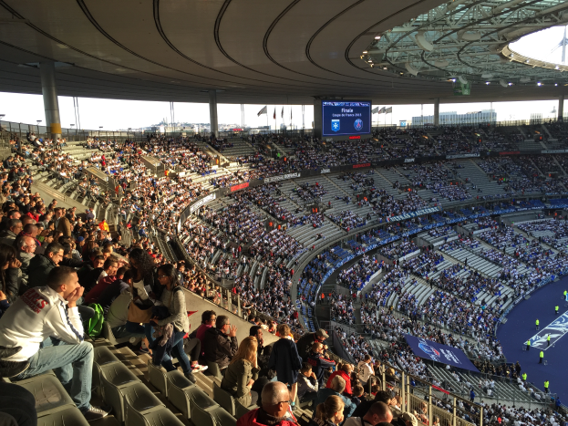 Eine große Menschenmenge sitzt im Allianz Stadion in München, Deutschland, und schaut ein Fußballspiel, mit einer Bühne auf der rechten Seite, Fahnen, Stangen und einem Bildschirm im Hintergrund und dem Himmel oben im Bild.