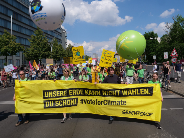 Eine Gruppe von Menschen geht eine Straße entlang und hält ein gelbes Banner mit der Aufschrift "Wähle für das Klima", mit Ballons, Schildern, Fahrrädern, Bäumen, Laternenpfählen und Gebäuden im Hintergrund unter einem klaren blauen Himmel.