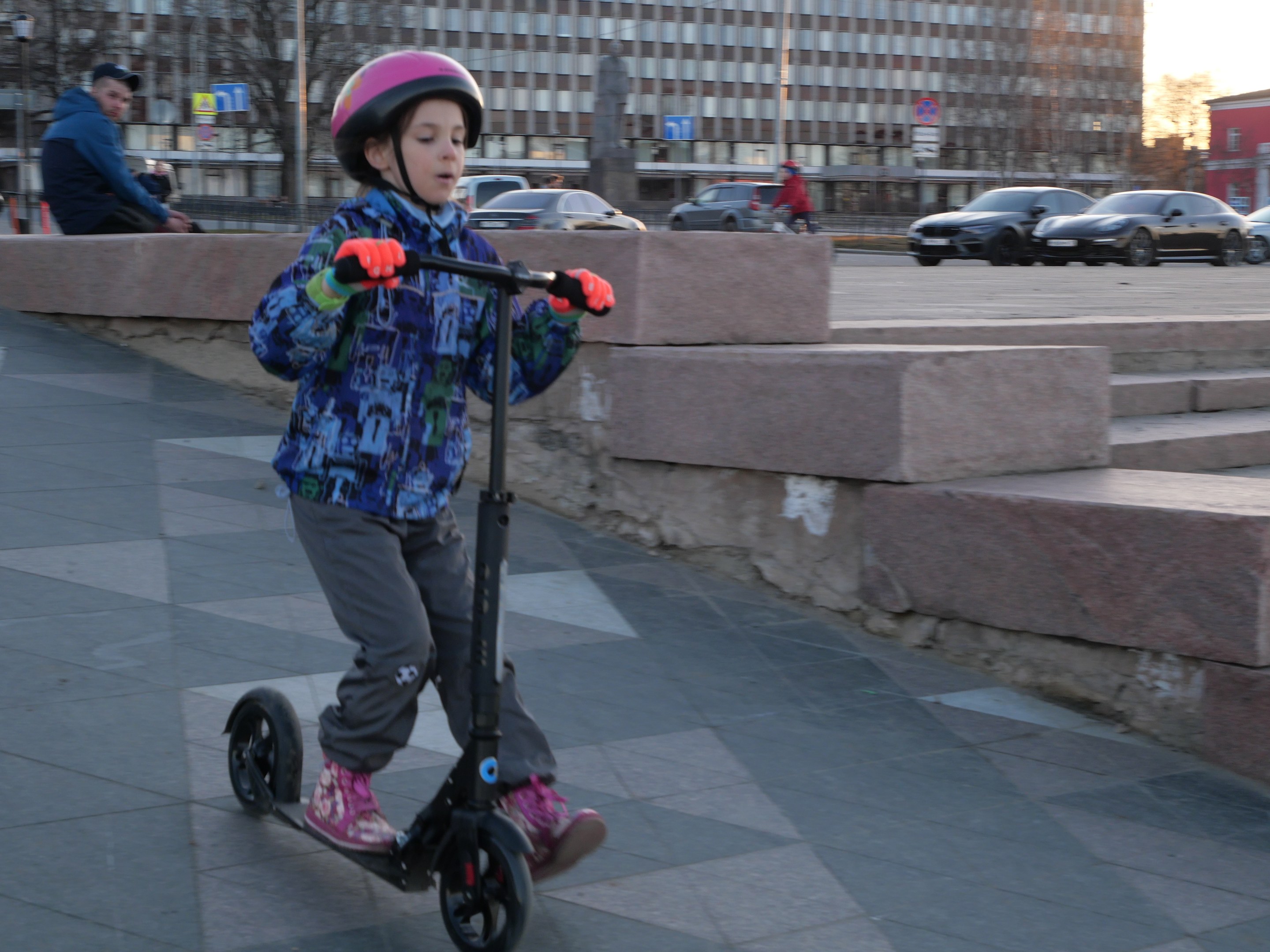 Ein Junge mit Helm und Handschuhen fährt auf einem Gehweg mit einem Roller, im Hintergrund sind Treppen, Fahrzeuge, Menschen, Bäume, Pfosten, Bretter, Gebäude und ein klarer blauer Himmel zu sehen.