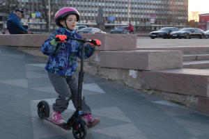 Ein Junge mit Helm und Handschuhen fährt auf einem Gehweg mit einem Roller, im Hintergrund sind Treppen, Fahrzeuge, Menschen, Bäume, Pfosten, Bretter, Gebäude und ein klarer blauer Himmel zu sehen.