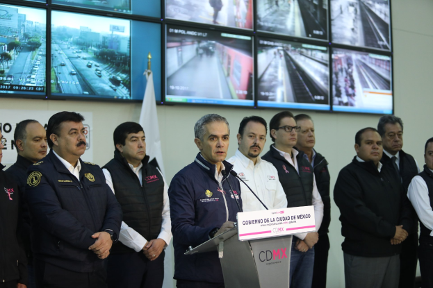 Männergruppe vor einem Podium mit Mikrofon und einer Tafel mit der Aufschrift "Mexikanische Zoll- und Grenzschutzbehörde", mit Fernsehgeräten und einer Flagge an der Wand dahinter.