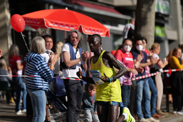 Ein Mann in einem gelben T-Shirt läuft durch eine jubelnde Menge während eines Marathons, mit einem Schirm und einem Kinderwagen im Hintergrund.