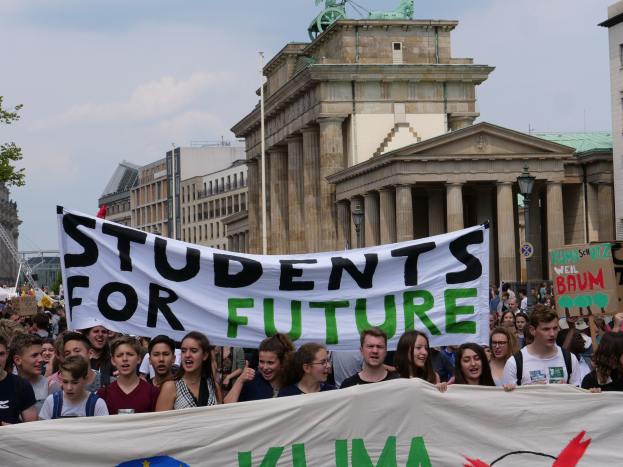 Gruppe von Studenten, die in Berlin mit einer bunt bemalten "Students for Future"-Schlagzeile vor Gebäuden, Bäumen und Himmel marschieren.