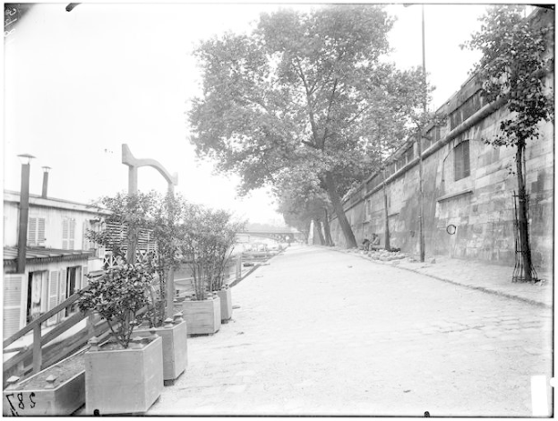 Ein Schwarz-Weiß-Foto einer Straße mit einer Wand auf der rechten Seite, Bäumen und Pflanzen auf der linken Seite und einer Brücke im Hintergrund, mit Text unten, der "New York City, New York, New Jersey, NY, USA, USA - Straßenansicht des New York State Penitentiaries" lautet.