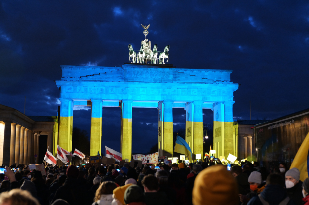 Eine Menschenmenge mit Fahnen und Plakaten steht vor dem Brandenburger Tor in Berlin, Deutschland, mit seinen Statuen und Säulen, die unter einem bewölkten Himmel zu sehen sind; ein Banner mit Text befindet sich auf der rechten Seite.