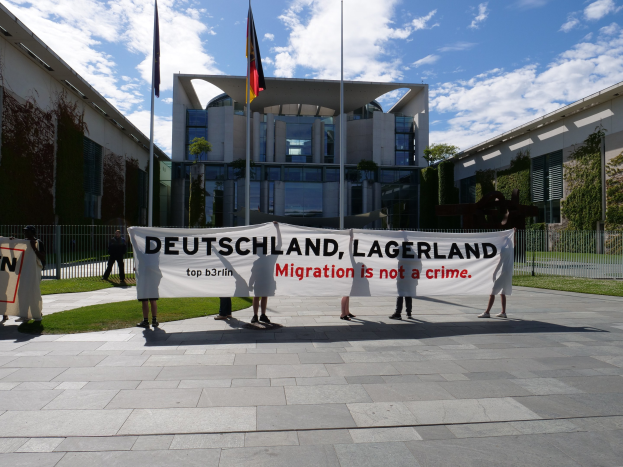 Gruppe von Menschen mit einem Banner, auf dem 'Deutschland, Lagerland Migration ist kein Verbrechen' steht, vor einem Gebäude mit Fenstern, umgeben von Gras, einem Metallzaun, Pflanzen, Bäumen und Fahnen mit Stangen unter einem bewölkten Himmel.