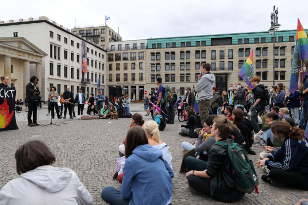 Eine Gruppe von Menschen, die auf dem Boden vor einer Menge sitzen, die Fahnen und Schilder hölt, während einer Anti-Schwulen-Demo in Berlin, mit einer Statue, Gebäuden und einem Mikrofonständer zu sehen.