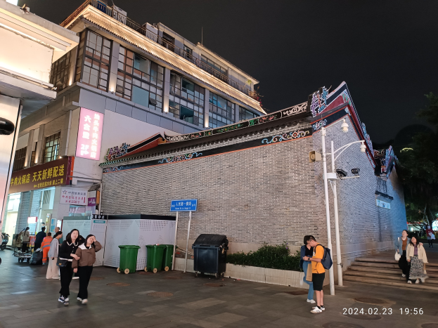 Menschen gehen nachts eine Straße entlang, beleuchtet von Straßenlaternen und umgeben von Gebäuden mit Fenstern, Schildern, Mülltonnen, Pflanzen, Bäumen und einem bewölkten Himmel, einige tragen Taschen.