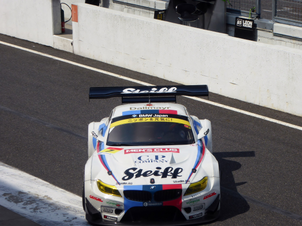 BMW M6 GT3 Rennauto auf einer Rennstrecke mit Text auf dem Fahrzeug, einer Wand, Maschendrahtzaun und weiteren Elementen im Hintergrund.