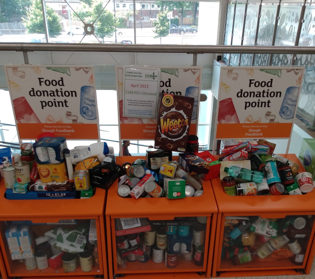 Orange Schränke mit Lebensmittelspenden gefüllt, darunter Flaschen, Dosen und Kartons, mit Informationsschildern in der Nähe und einem Blick auf Fahrzeuge, Bäume, Gebäude und Himmel durch Glasfenster.