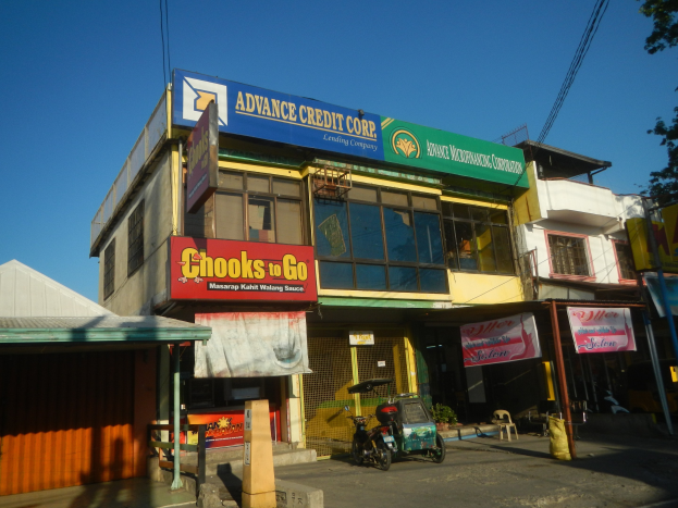 Ein Gebäude mit einer Tafel, auf der "Advance Credit Corp" steht, an einer Straße mit Fahrzeugen, Strommasten, Bäumen und Werbetafeln unter einem klaren blauen Himmel.