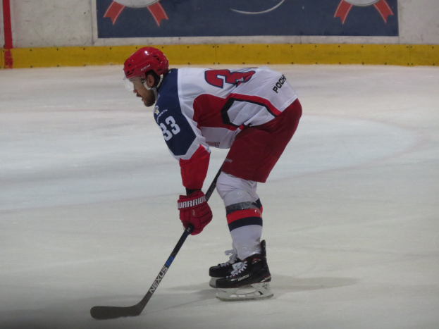 Eishockey-Spieler in voller Ausrüstung auf dem Eis mit einem Banner mit Text und Logos im Hintergrund.