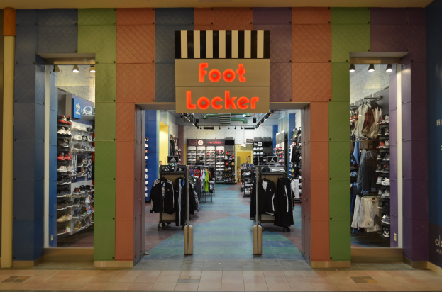 Innenansicht eines Foot Locker-Schuhgeschäfts mit Kleidung an Bügeln, Waren auf Gestellen und beleuchteter Beleuchtung; der Boden ist unten sichtbar.