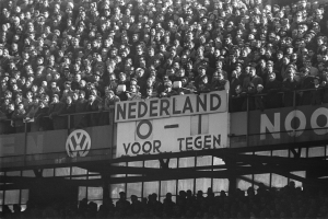 Ein Schwarz-Weiß-Foto einer großen Menschenmenge in einem Stadion, mit einer Tafel in der Mitte und ein paar Individuen am unteren Rand.