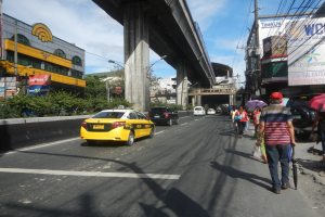 Ein gelbes Taxi fährt auf einer Stadtstraße mit hohen Gebäuden, Fußgängern mit Regenschirmen, einer Brücke im Hintergrund, bewölktem Himmel und Strommasten mit Überlandleitungen.