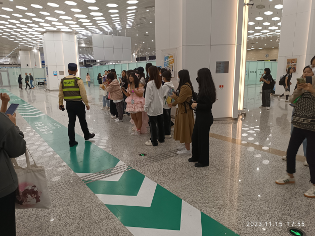 Menschen in einer Schlange an einem Flughafen-Sicherheits-Check-In-Bereich mit Taschen und Mobiltelefonen, mit Säulen und Deckenlampen im Hintergrund.
