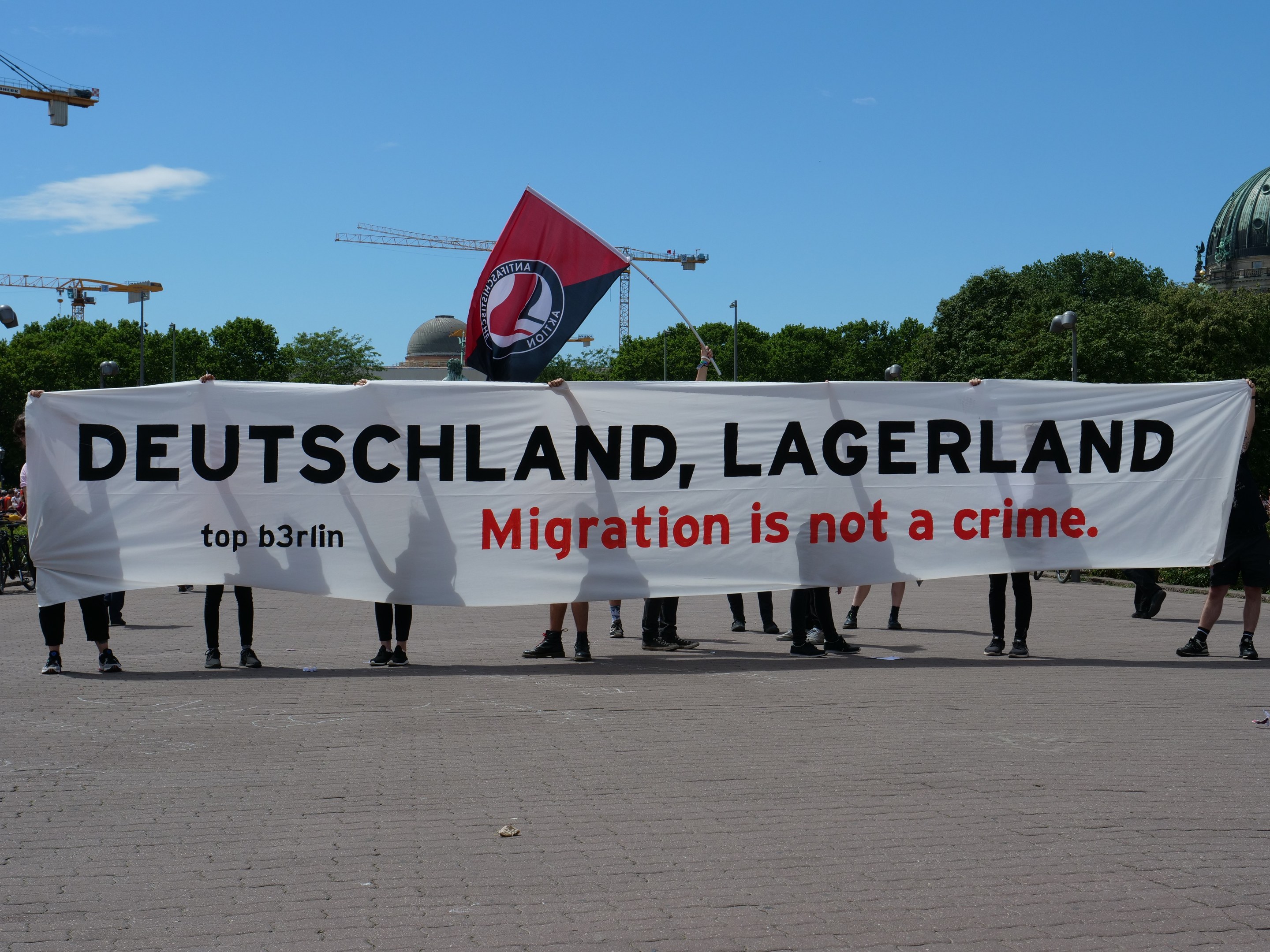 Eine Gruppe von Menschen hält ein Transparent mit der Aufschrift 'Deutschland, Lagerland Migration ist kein Verbrechen' und eine Flagge, mit Straßenlaternen, Bäumen, Kränen, einem Gebäude und einem bewölkten Himmel im Hintergrund.